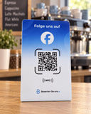 Blau-weißer Facebook-Aufsteller mit NFC-Symbol und QR-Code auf einem Holztresen, im Hintergrund eine Kaffeemaschine.