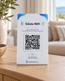 Weiß-blauer Gäste-WLAN-Aufsteller mit QR-Code auf einer Holzkommode, dahinter ein unscharfer Wohnbereich mit Sofa.